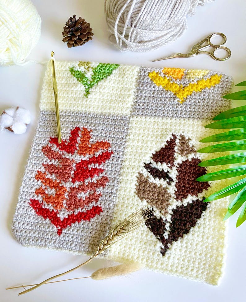 May include: A square, crocheted piece with four leaf designs in shades of red, orange, yellow, green, and brown. The background is a mix of cream and gray. A gold crochet hook, scissors, and yarn are nearby.