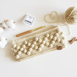 May include: A beige crocheted headband with a textured bobble stitch pattern. The headband is laid flat on a white surface with a wooden crochet hook, a ball of yarn, and two wooden tags that say "Love is Kind".
