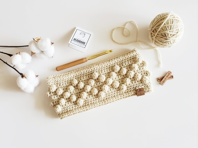 May include: A beige crocheted headband with a textured bobble stitch pattern. The headband is laid flat on a white surface with a wooden crochet hook, a ball of yarn, and two wooden tags that say "Love is Kind".