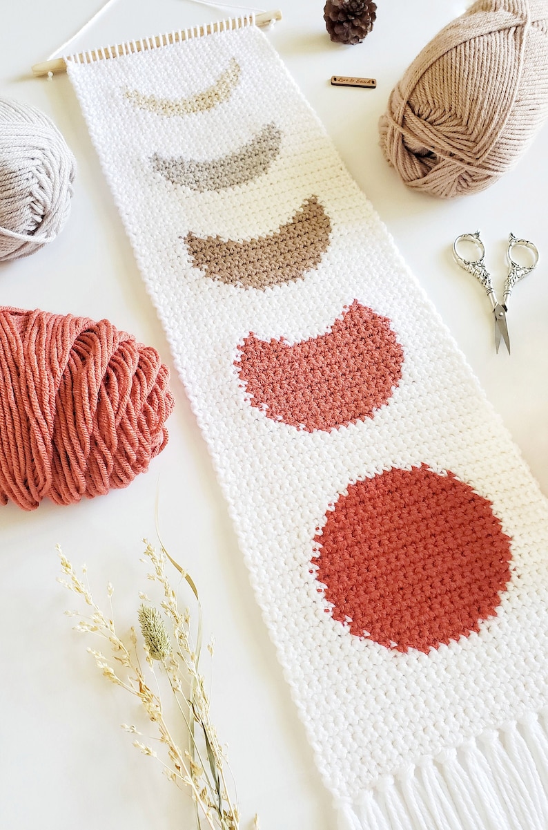 May include: A white macrame wall hanging with a fringe at the bottom. The wall hanging features five crescent moon shapes in shades of beige, brown, and orange.
