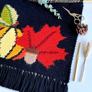 May include: A black, handmade wall hanging with a fall theme. It features a large red maple leaf, a yellow and green gourd, and a brown acorn, all in a cross-stitch style. The bottom has a fringe. A wooden dowel and gold scissors are also visible.