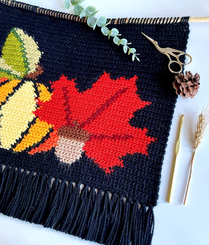 May include: A black, handmade wall hanging with a fall theme. It features a large red maple leaf, a yellow and green gourd, and a brown acorn, all in a cross-stitch style. The bottom has a fringe. A wooden dowel and gold scissors are also visible.