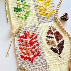 May include: A handmade crochet wall hanging featuring four leaf designs in green, yellow, red, and brown. The leaves are set against a cream and gray background. A wooden dowel and fringe complete the piece. A gold crochet hook and scissors are also visible.