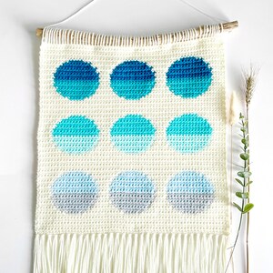 May include: A white crocheted wall hanging with a fringe border. The hanging features nine ombre circles in shades of blue and gray.