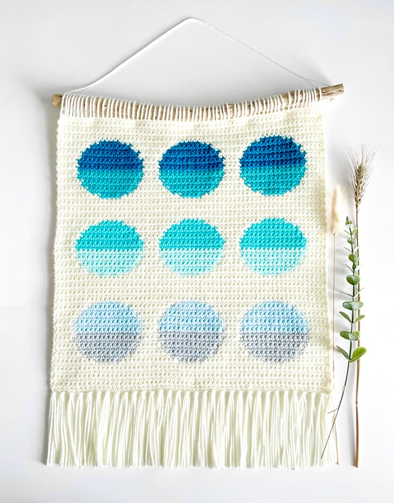 May include: A white crocheted wall hanging with a fringe border. The hanging features nine ombre circles in shades of blue and gray.