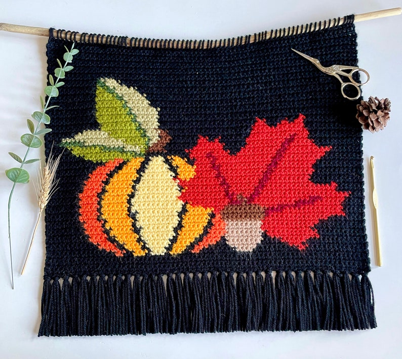 May include: Black crochet wall hanging with a fall theme. Features a pumpkin, maple leaf, and acorn in orange, yellow, green, and red. The bottom has a fringe. A wooden dowel and scissors are at the top.