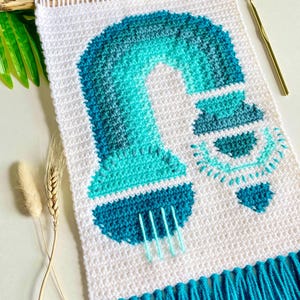 Puede incluir: Un tapiz de pared blanco tejido a ganchillo con un diseño turquesa y aguamarina que representa una lámpara. El diseño está sujeto a una clavija de madera y tiene una borla turquesa en la parte inferior. Se ve un ganchillo dorado.