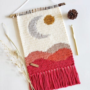 May include: A white crocheted wall hanging with a gray crescent moon, a yellow sun, and red, orange, and brown mountains. The hanging has a fringe of red yarn and a brown leather tag with the text "Yarn & Stitch".