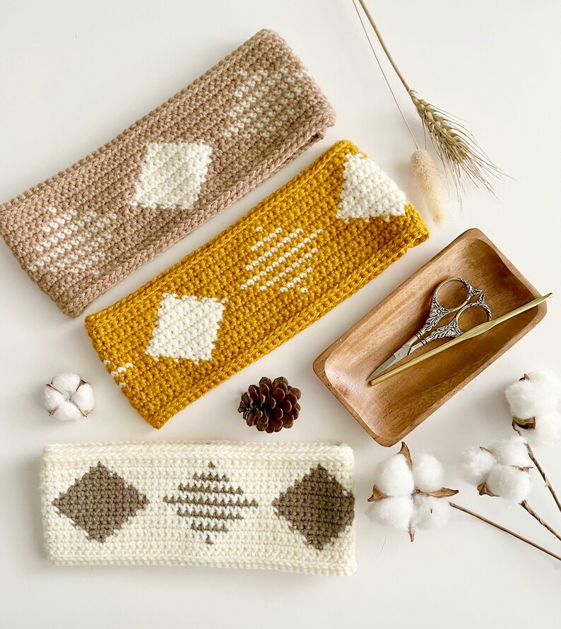 Pode incluir: Tr&ecirc;s faixas de cabe&ccedil;a de croch&ecirc; em diferentes cores e padr&otilde;es. As faixas de cabe&ccedil;a s&atilde;o feitas com fio branco, marrom e amarelo. As faixas de cabe&ccedil;a est&atilde;o dispostas em uma superf&iacute;cie branca com bolas de algod&atilde;o e uma bandeja de madeira com tesoura.