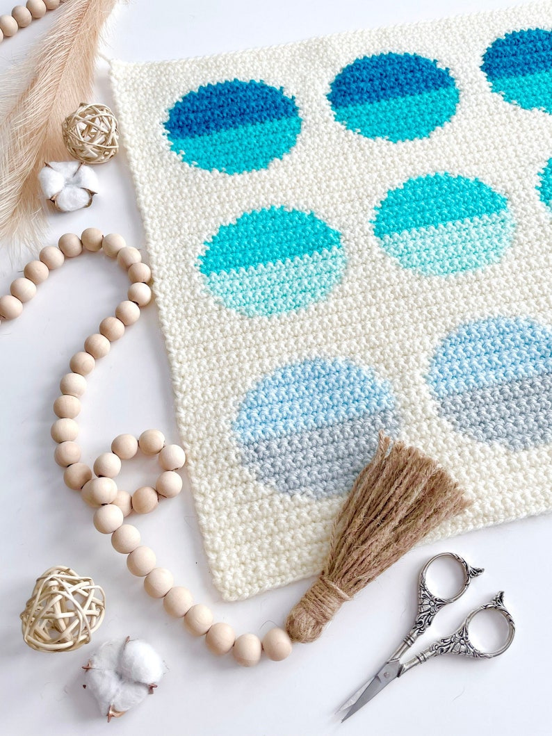 May include: A white crocheted wall hanging with a geometric pattern of blue and gray circles. The hanging has a brown tassel attached to the bottom.
