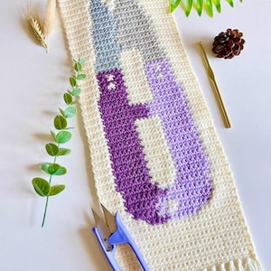 Puede incluir: Un tapiz rectangular de ganchillo en color crema con un diseño gráfico de dos tijeras con hojas grises y mangos morados. La pieza está suspendida de un palo de madera y terminada con flecos en el borde inferior.