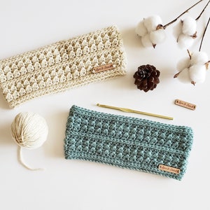 Peut inclure: Deux bandeaux en crochet, l'un beige et l'autre turquoise, avec une étiquette en cuir marron qui dit "Love & Stitch". Les bandeaux sont sur une surface blanche avec un écheveau de laine, un crochet et des boules de coton.