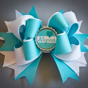 May include: A teal and white hair bow with a silver center piece that says "Alyssa" with a heart.
