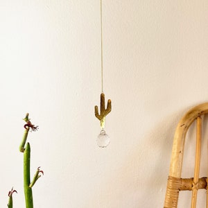 May include: A gold metal cactus hanging ornament with a clear crystal prism hanging from the bottom. The ornament is hanging from a gold chain.