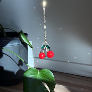 May include: A cherry-shaped charm with two red orbs and green accents, suspended from a gold-colored chain. The charm is held up in front of a green plant with large leaves, with sunlight creating a bright background.