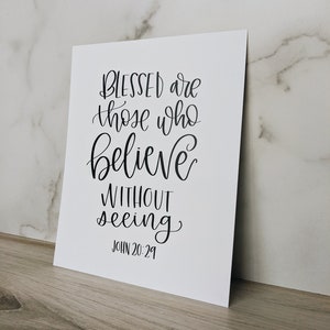 May include: A white print with black calligraphy. The text reads "BLESSED are those who believe WITHOUT seeing" and "JOHN 20:29". The print is displayed on a wooden surface with a marble background.