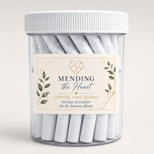 Op de afbeelding: Een doorzichtige plastic pot met een wit geribbeld deksel, gevuld met strak opgerolde witte papieren briefjes. Het etiket bevat een gouden gebarsten hart-icoon, groene bladillustraties en de tekst MENDING the Heart COMFORT HOPE HEALING 60 Days of Comfort for the Journey Ahead.