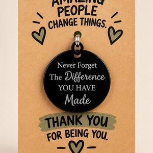 May include: A round black metal pendant attached to a rectangular tan card. The pendant is engraved with the text: Never Forget The Difference YOU HAVE Made. The card features the text: AMAZING PEOPLE CHANGE THINGS. THANK YOU FOR BEING YOU.
