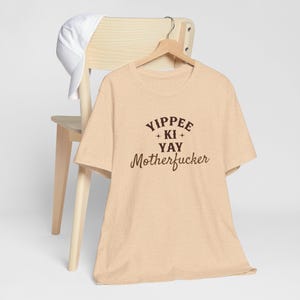 May include: A beige t-shirt with the text "Yippee + Ki + Yay Motherfucker" printed in brown on the front.