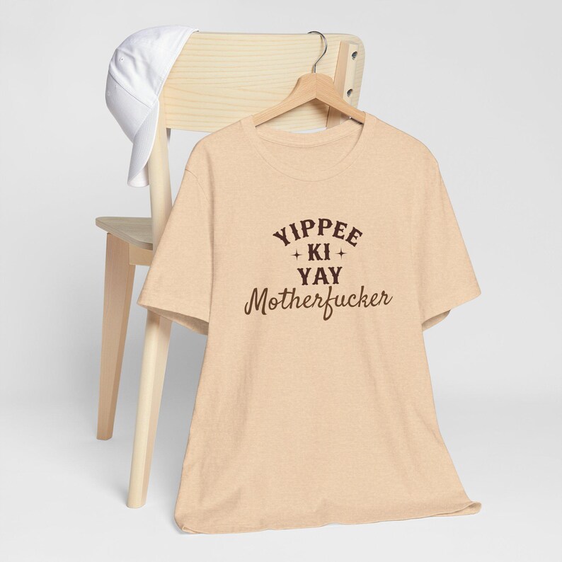 May include: A beige t-shirt with the text "Yippee + Ki + Yay Motherfucker" printed in brown on the front.
