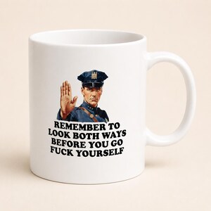 May include: A white ceramic mug featuring a graphic of a police officer in a blue uniform holding up a hand in a stop gesture. Below the illustration, the text reads: REMEMBER TO LOOK BOTH WAYS BEFORE YOU GO FUCK YOURSELF in bold black letters.