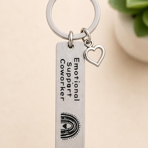 May include: A silver-coloured metal keyring featuring a rectangular pendant engraved with the text Emotional Support Coworker and a graphic of a rainbow. The keyring includes a circular split ring and a small, open heart-shaped charm attached.
