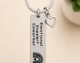 May include: A silver-coloured metal keyring featuring a rectangular pendant engraved with the text Emotional Support Coworker and a graphic of a rainbow. The keyring includes a circular split ring and a small, open heart-shaped charm attached.