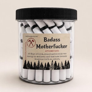 May include: A clear plastic jar with a black screw-top lid filled with white paper scrolls tied with black bands. The label reads Badass Motherfucker Affirmations, 60 Days of bold, unapologetic kick-ass energy to motivate and dominate because weakness isn't an option.