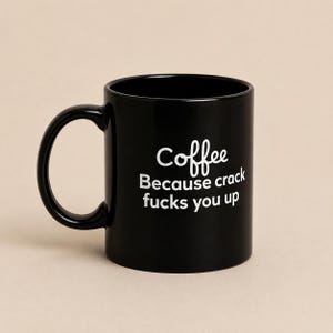 May include: A black ceramic mug with a rounded handle and white printed text that reads: Coffee Because crack fucks you up. The mug has a smooth, glossy finish and a standard cylindrical shape, designed for holding hot or cold beverages.
