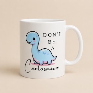 Op de afbeelding: Een witte keramische mok met een cartoonillustratie van een lichtblauwe dinosaurus met roze wangen. De tekst op de mok luidt DON'T BE A Cuntasaurus in een combinatie van dikke blokletters en een zwart cursief lettertype.
