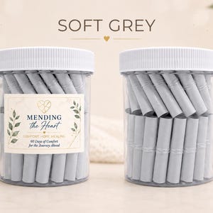 May include: Two clear plastic jars with white lids, each filled with rolled-up light grey paper slips. The left jar features a label reading MENDING the Heart COMFORT HOPE HEALING 60 Days of Comfort for the Journey Ahead with leaf graphics.