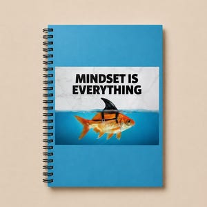 May include: A blue spiral-bound notebook featuring a graphic of a goldfish wearing a black shark fin harness. Above the fish, the text "MINDSET IS EVERYTHING" is printed in bold black letters against a white marble-textured background.