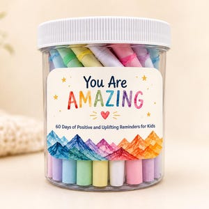Könnte beinhalten: Ein durchsichtiges Kunststoffglas mit weißem Deckel, gefüllt mit pastellfarbenen Papierrollen. Das Etikett zeigt den Text "You Are AMAZING" und "60 Days of Positive and Uplifting Reminders for Kids" über einer bunten Aquarell-Berglandschaft.