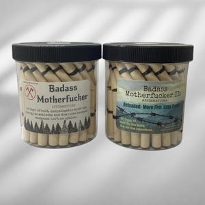 May include: Two clear plastic jars with black lids filled with rolled paper. The labels read "Badass Motherfucker" and "Badass Motherfucker II: Reloaded." The labels include text about affirmations and motivation.