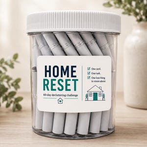 May include: A clear plastic jar with a white ribbed lid filled with rolled-up gray paper slips. The label reads HOME RESET 60-day decluttering challenge, with a house icon and text stating One card, One task, One less thing to stress about.