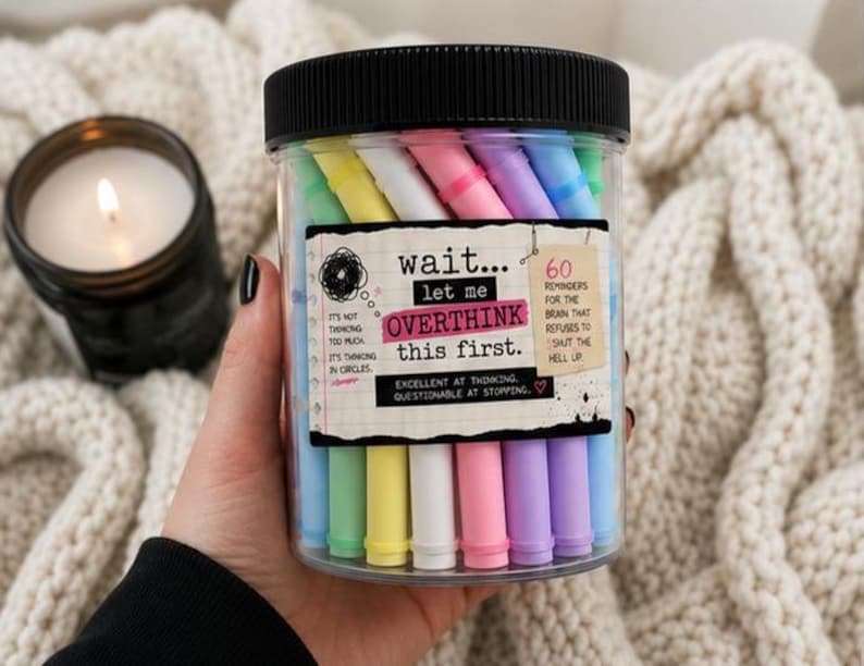 May include: A clear plastic jar with a black lid containing pastel-colored markers. The label reads: wait... let me OVERTHINK this first. 60 REMINDERS FOR THE BRAIN THAT REFUSES TO SHUT THE HELL UP. IT'S NOT THINKING TOO MUCH, IT'S THINKING IN CIRCLES.