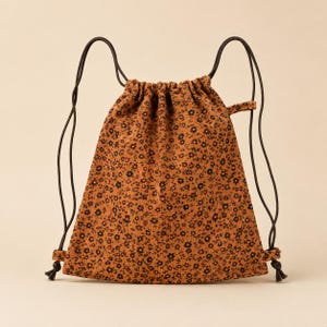 May include: A drawstring backpack made of brown corduroy fabric featuring a repeating pattern of small black flowers with white centres. The bag includes thin dark brown cord straps that cinch the top opening and are knotted at the bottom corners.