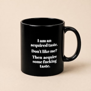 May include: A glossy black ceramic mug featuring white printed text that reads: I am an acquired taste. Don't like me? Then acquire some fucking taste. The mug has a standard cylindrical shape with a curved handle on the right side and a smooth finish.