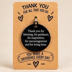 May include: A round black metal pendant attached to a tan card. The card reads THANK YOU FOR ALL THAT YOU DO and YOU MAKE A DIFFERENCE EVERY DAY. The pendant is engraved with the text Thank you for listening, for guidance, for inspiration, for encouragement and for being here.
