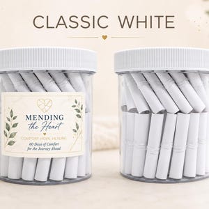 May include: Two clear plastic jars with white ribbed lids, each filled with numerous tightly rolled white paper scrolls. The left jar features a label with the text MENDING the Heart, COMFORT-HOPE-HEALING, 60 Days of Comfort for the Journey Ahead.