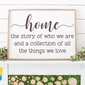 May include: A framed wooden sign with the word "home" in a cursive font. The text below reads "the story of who we are and a collection of all the things we love." The sign is displayed on a white mantel with greenery.