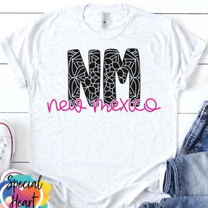 May include: A white t-shirt with a black graphic of the letters "NM" with a mandala design inside each letter. The words "new mexico" are written in pink cursive below the letters.