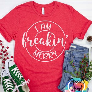 May include: Red t-shirt with white text that reads "I am freakin' merry" in a circle. The shirt is laid flat on a white surface with a pair of green converse shoes, blue jeans, and a sprig of red berries.
