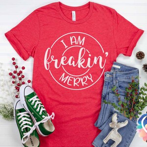 May include: Red t-shirt with white text that reads "I am freakin' merry" in a circle. The shirt is laid flat on a white surface with a pair of green converse shoes, blue jeans, and a white reindeer figurine.