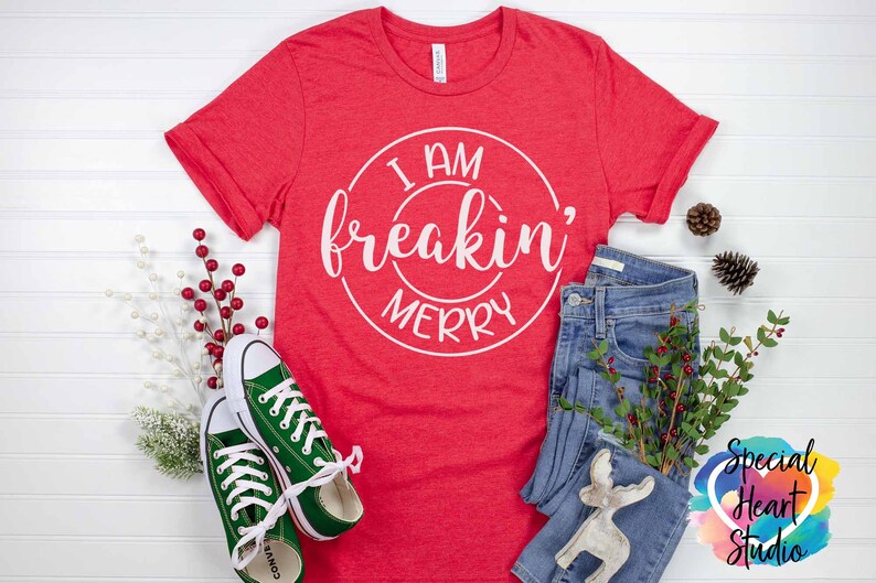 May include: Red t-shirt with white text that reads "I am freakin' merry" in a circle. The shirt is laid flat on a white surface with a pair of green converse shoes, blue jeans, and a white reindeer figurine.