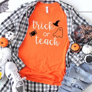 以下が含まれることがあります： 白い文字で「trick or teach」と書かれたオレンジ色のTシャツ。テキストの上に黒いクモと魔女の帽子をかぶった幽霊がいます。Tシャツは、白木の表面に黒と白の格子縞のシャツ、青いジーンズ、ハロウィンの装飾と一緒に置かれています。