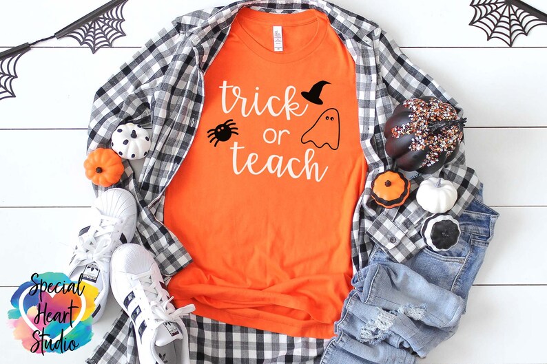 以下が含まれることがあります： 白い文字で「trick or teach」と書かれたオレンジ色のTシャツ。テキストの上に黒いクモと魔女の帽子をかぶった幽霊がいます。Tシャツは、白木の表面に黒と白の格子縞のシャツ、青いジーンズ、ハロウィンの装飾と一緒に置かれています。