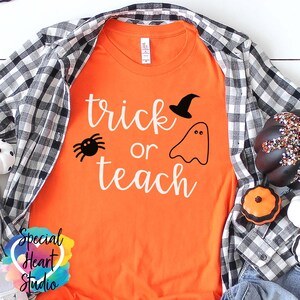 以下が含まれることがあります： オレンジ色のTシャツに白い文字で「trick or teach」と書かれています。文字の上に黒いクモ、幽霊、魔女の帽子があります。