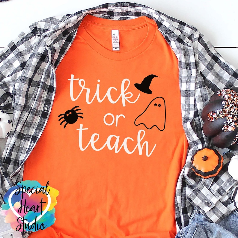 以下が含まれることがあります： オレンジ色のTシャツに白い文字で「trick or teach」と書かれています。文字の上に黒いクモ、幽霊、魔女の帽子があります。