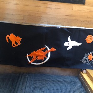 May include: Black fabric Halloween table runner with orange and white felt appliques of a cat, ghost, witch, pumpkin, and spider web.